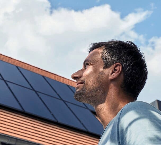 Un hombre de pie frente a una casa con paneles fotovoltaicos en el tejado